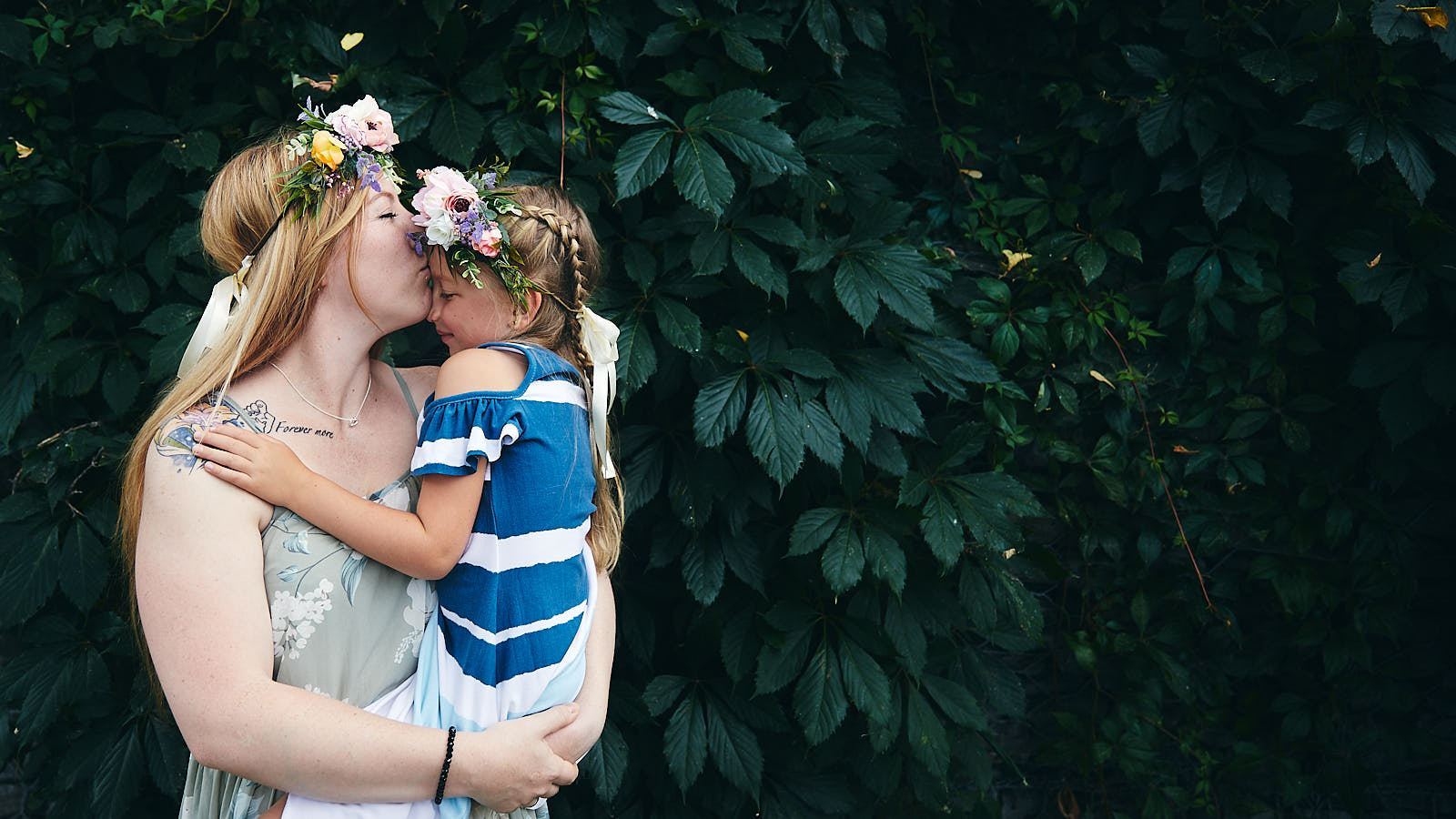 Mère et fille s'embrassent avec fleurs dans les cheveux devant un mur de feuilles.