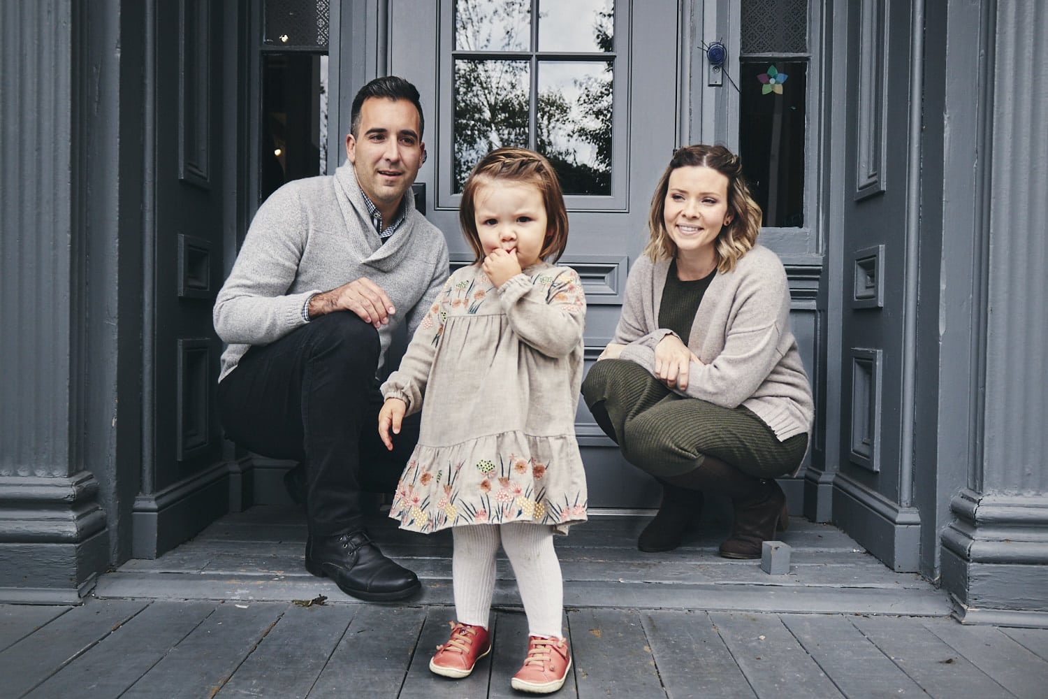 Famille souriante avec une petite fille devant une porte en bois gris foncé.